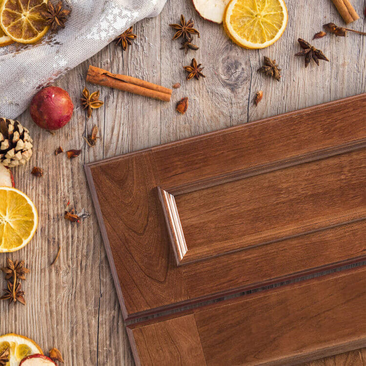 A flat panel cabinet door style with a delicious, red-orange stained finish on cherry wood. Cinnamon sticks and cider supplies are scattered around the door on a table to emphasize the warm stain color.