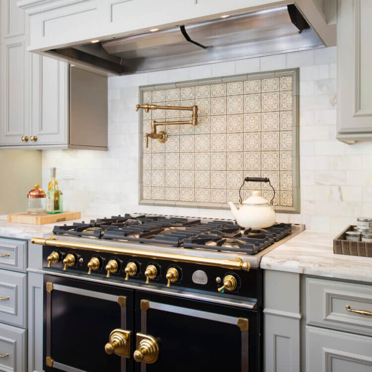 Beautiful slide-in range with double ovens in a gray kitchen design with gray painted kitchen cabinets and light gray backsplash tiles. Gold hardware, pot filler, and accents add warmth to the cool color palette. A detailed wood hood adds elegance to the design.