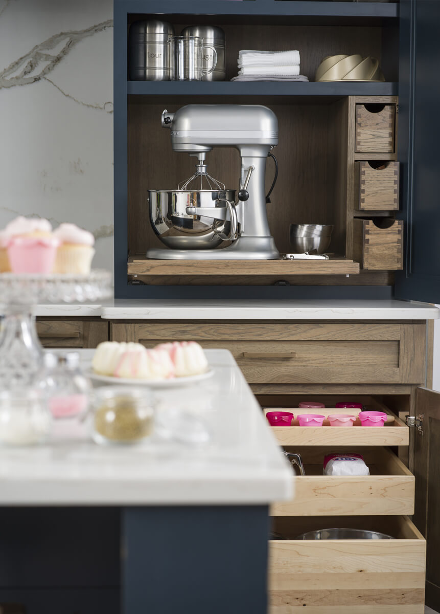 A baking center larder cabinet with pantry storage and organized roll-out shelves for baking supplies and tools. A flat roll-out shelf holds a kitchen aid mixer and makes it easy to hide it away behind cabinet doors in this navy blue and hickory wood kitchen.