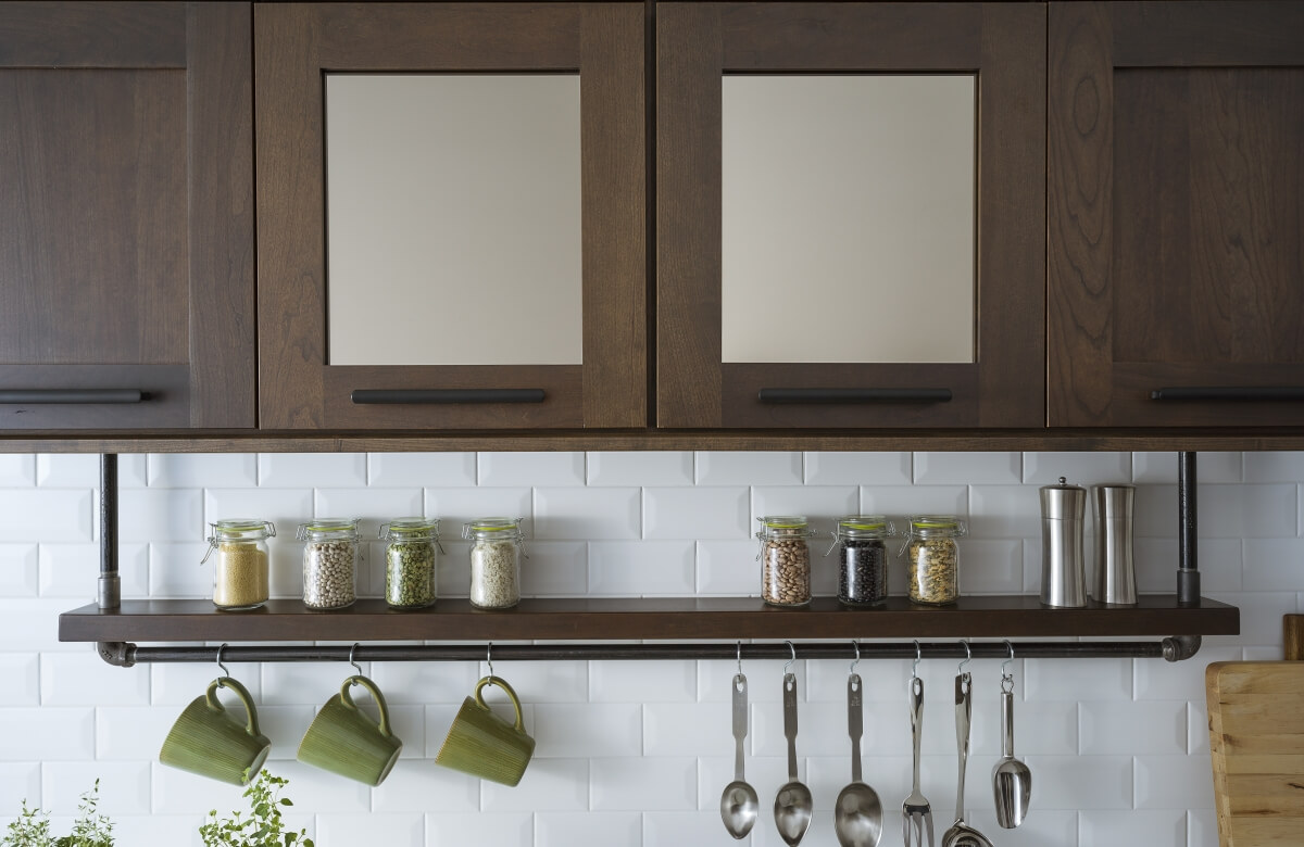 A floating shelf below the wall cabinets creates an industrial style by using basic pipe fixtures to make it appear like the shelving is hanging by pipes from the wall cabinets. The shelf provides a place o display everyday used supplies like spices, coffee cups, and cooking utensils. Mirrored glass cabinets above conceal what's behind the cabinet doors but give the appearance of a glass door.