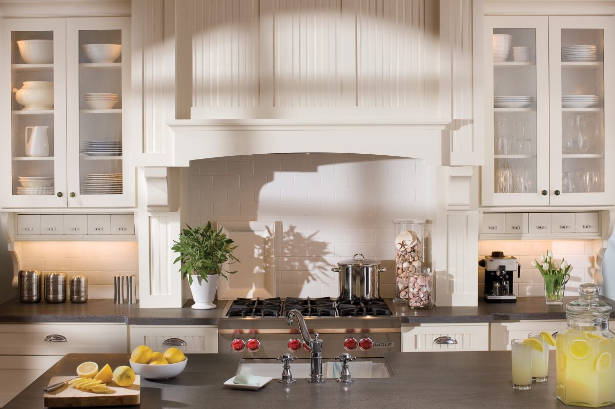 A beautiful example of a cottage style kitchen with white painted kitchen cabinets, a wood hood, kitchen island, and white subway tile backsplash. Cottage cabinets by Dura Supreme Cabinetry.