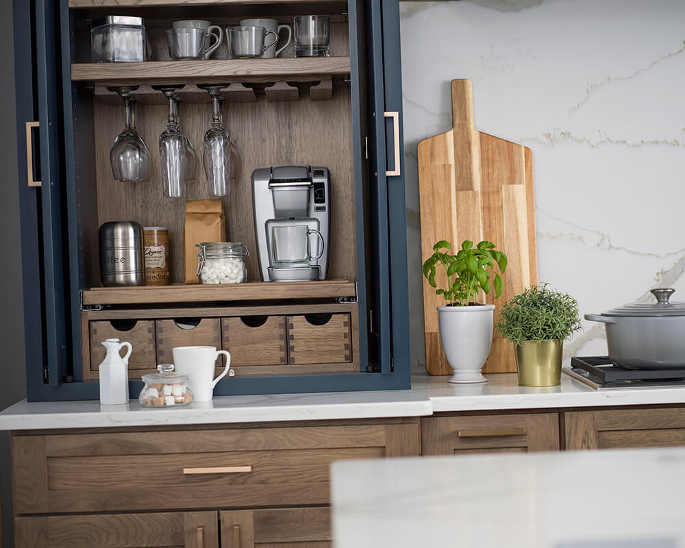 This breakfast work zone is design has everything the homeowner needs for crafting beverages for all occasions as well as a place for preparing breakfast. The larder cabinet houses a powered coffee machine, with coffee cups and wine glasses above and all the supplies for making a wide variety of beverages in the cubbies below. The apothecary drawers below hold stir sticks, tea bags, hot cocoa packets, sugar, sweeteners, and liquid creamer cups.