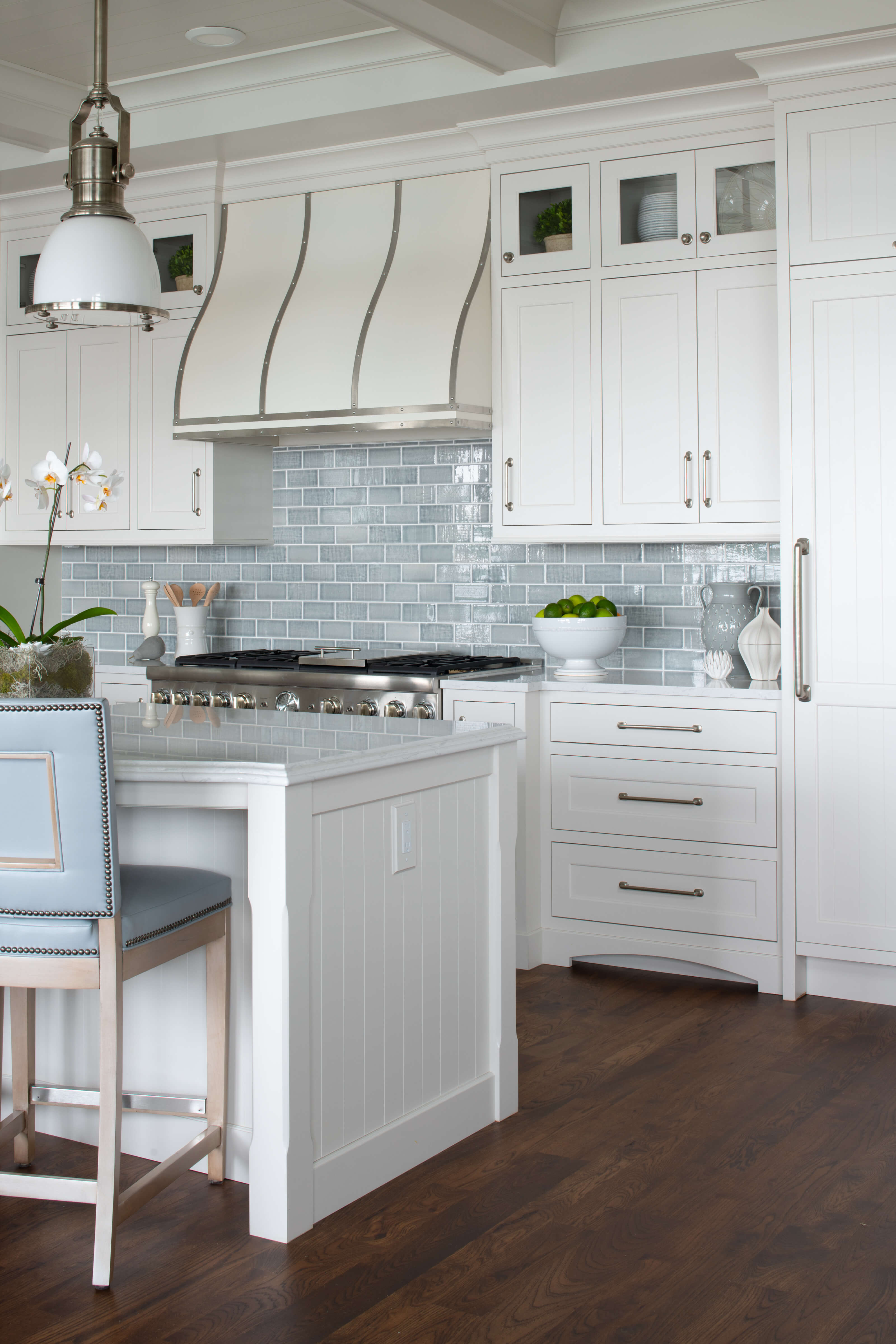 A coastal style kitchen with white painted inset cabinets with shiplap v-groove paneled doors and kitchen island end cap. The design features a light blue backsplash an island stools.