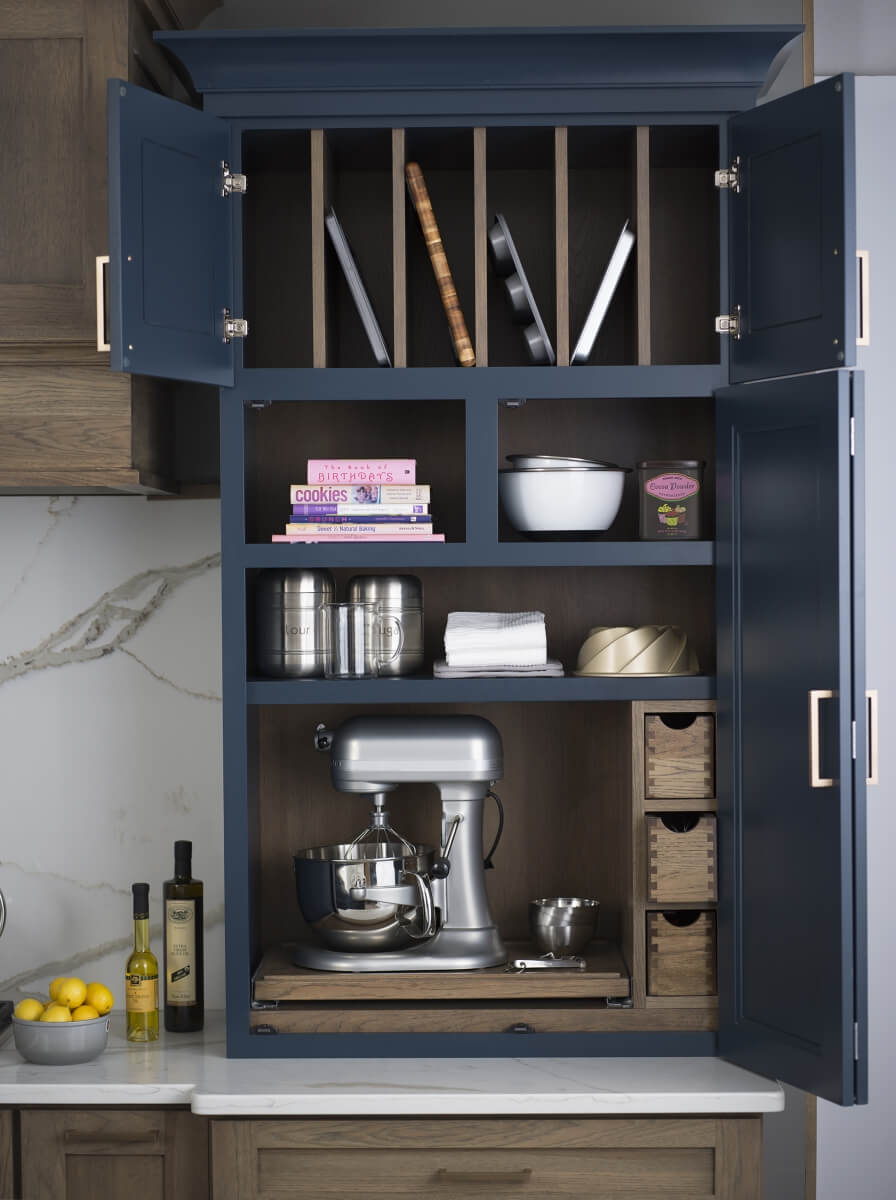 A baking center larder cabinet with pantry storage and organized roll-out shelves for baking supplies and tools. A flat roll-out shelf holds a kitchen aid mixer and makes it easy to hide it away behind cabinet doors in this navy blue and hickory wood kitchen.
