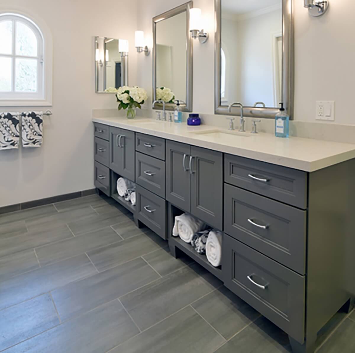 A master bathroom with a dark gray painted vanity with open storage for bath towels. Featuring a nickel finish on the cabinet hardware, plumbing fixtures, faucet, light fixtures, and bathroom mirrors frames. A lighter gray colored floor tile complements the dark gray vanity cabinets.