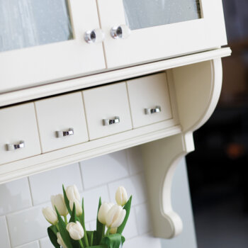 A cottage style kitchen with detailed corbels, antique glass, off-white paint, white subway tiles, and a row of adorable apothecary drawers below the wall cabinet.