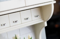 A cottage style kitchen with detailed corbels, antique glass, off-white paint, white subway tiles, and a row of adorable apothecary drawers below the wall cabinet.