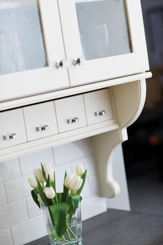 A cottage style kitchen with detailed corbels, antique glass, off-white paint, white subway tiles, and a row of adorable apothecary drawers below the wall cabinet.