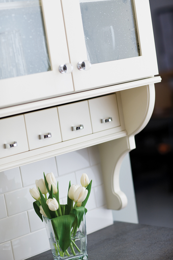 A cottage style kitchen with detailed corbels, antique glass, off-white paint, white subway tiles, and a row of adorable apothecary drawers below the wall cabinet.