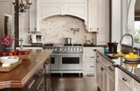 A traditional white kitchen with a grand, elegant wood hood over a double-wide range.