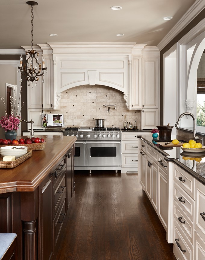 A traditional white kitchen with a grand, elegant wood hood over a double-wide range.