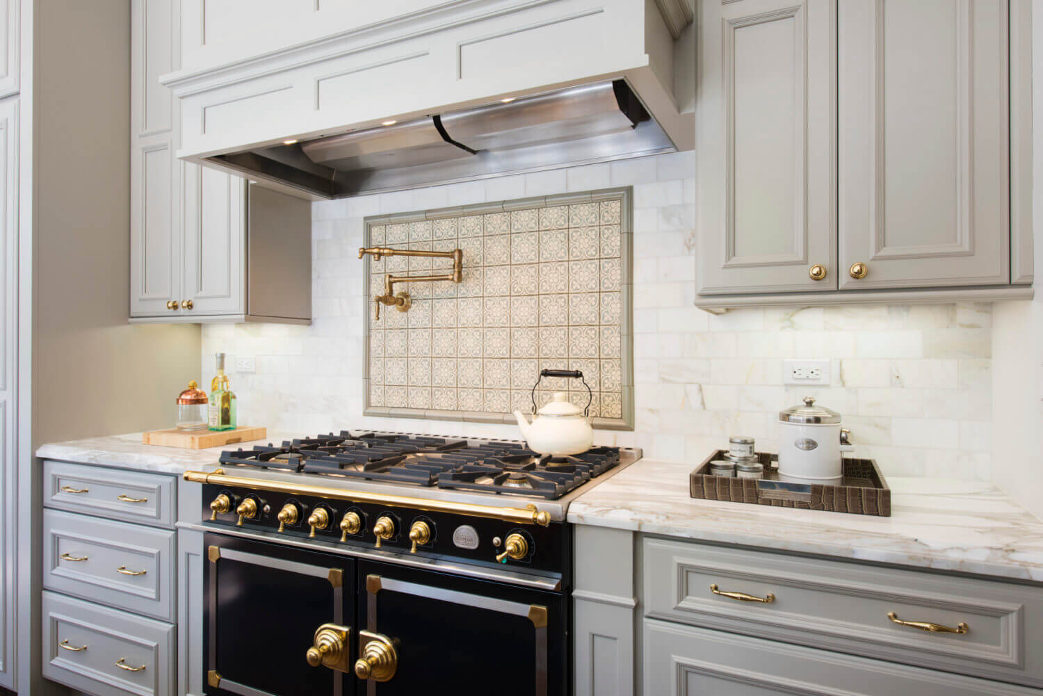 Beautiful slide-in range with double ovens in a gray kitchen design with gray painted kitchen cabinets and light gray backsplash tiles. Gold hardware, pot filler, and accents add warmth to the cool color palette. A detailed wood hood adds elegance to the design.