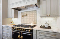 Beautiful slide-in range with double ovens in a gray kitchen design with gray painted kitchen cabinets and light gray backsplash tiles. Gold hardware, pot filler, and accents add warmth to the cool color palette. A detailed wood hood adds elegance to the design.