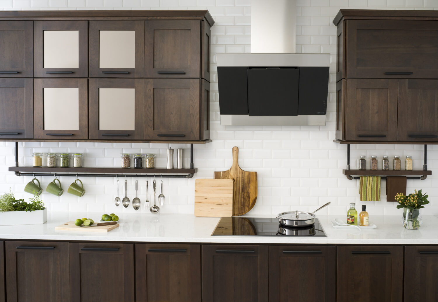 A floating shelf below the wall cabinets creates an industrial style by using basic pipe fixtures to make it appear like the shelving is hanging by pipes from the wall cabinets. The shelf provides a place o display everyday used supplies like spices, coffee cups, and cooking utensils. Mirrored glass cabinets above conceal what's behind the cabinet doors but give the appearance of a glass door. The dark stained cherry cabinets have a flat panel door style with full overlay construction.