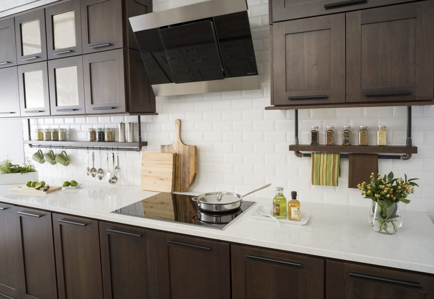 A floating shelf below the wall cabinets creates an industrial style by using basic pipe fixtures to make it appear like the shelving is hanging by pipes from the wall cabinets. The shelf provides a place o display everyday used supplies like spices, coffee cups, and cooking utensils. Mirrored glass cabinets above conceal what's behind the cabinet doors but give the appearance of a glass door. The dark stained cherry cabinets have a flat panel door style with full overlay construction.