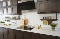 A floating shelf below the wall cabinets creates an industrial style by using basic pipe fixtures to make it appear like the shelving is hanging by pipes from the wall cabinets. The shelf provides a place o display everyday used supplies like spices, coffee cups, and cooking utensils. Mirrored glass cabinets above conceal what's behind the cabinet doors but give the appearance of a glass door. The dark stained cherry cabinets have a flat panel door style with full overlay construction.