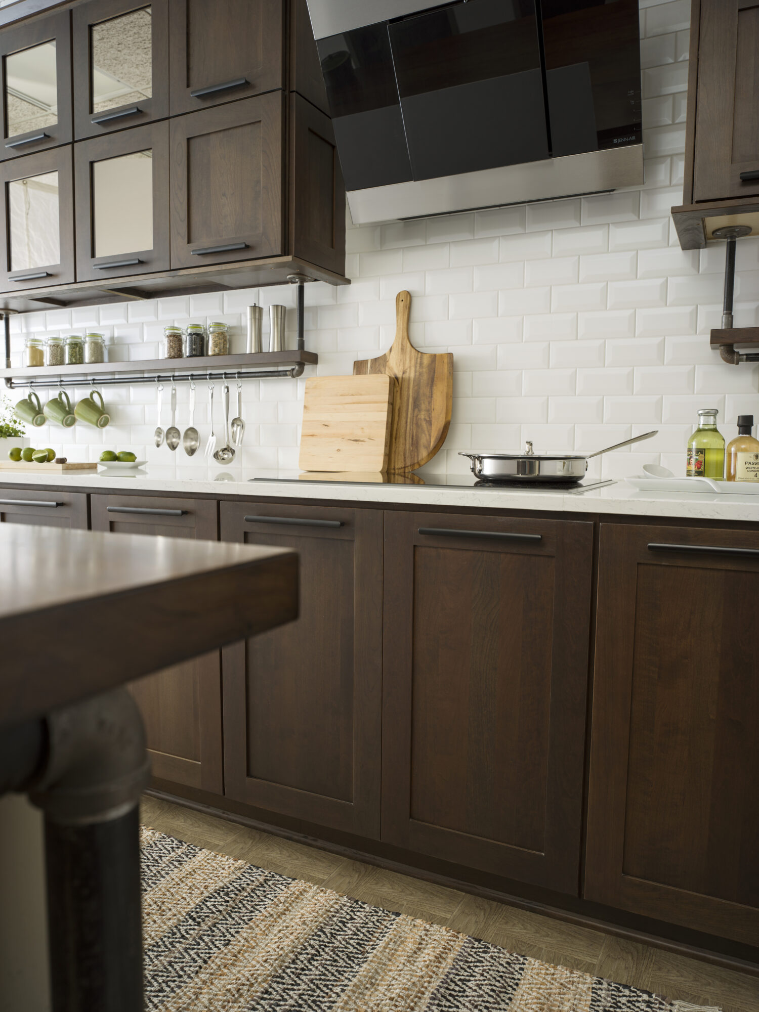 Cherry kitchen cabinets with a dark brown stain color with bright white countertops in a modern industrial style kitchen remodel. Industrial pipes are used to create the look of hanging floating shelves below the wall cabinets that displays everyday used supplies like coffee cups, utensils, spices, etc. The wall cabinets have mirror cabinet doors with a bronze-colored mirror insert.