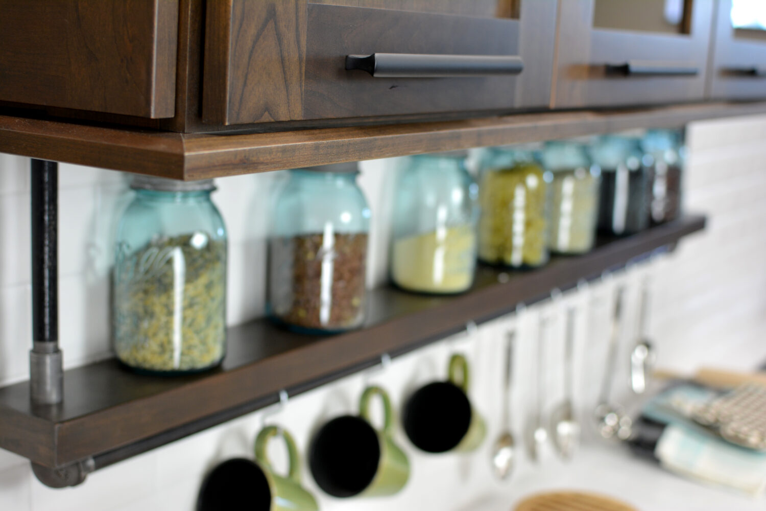 A floating shelf below the wall cabinets creates an industrial style by using basic pipe fixtures to make it appear like the shelving is hanging by pipes from the wall cabinets. The shelf provides a place o display everyday used supplies like spices, coffee cups, and cooking utensils.