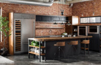 An Industrial style kitchen design in an urban loft with exposed brick walls and duct-work. The cabinets use a black painted finish and accents in a wood with a warm stain.