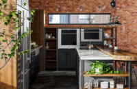 An Industrial style kitchen design in an urban loft with exposed brick walls and ductwork. The modern and rustic cabinets use a black painted finish and accents in a wood with a warm stain. Open shelves and black metal fixtures add to the style.