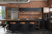 An Industrial style kitchen design in an urban loft with exposed brick walls and ductwork. The modern and rustic cabinets use a black painted finish and accents in a wood with a warm stain. Open shelves and black metal fixtures add to the style.