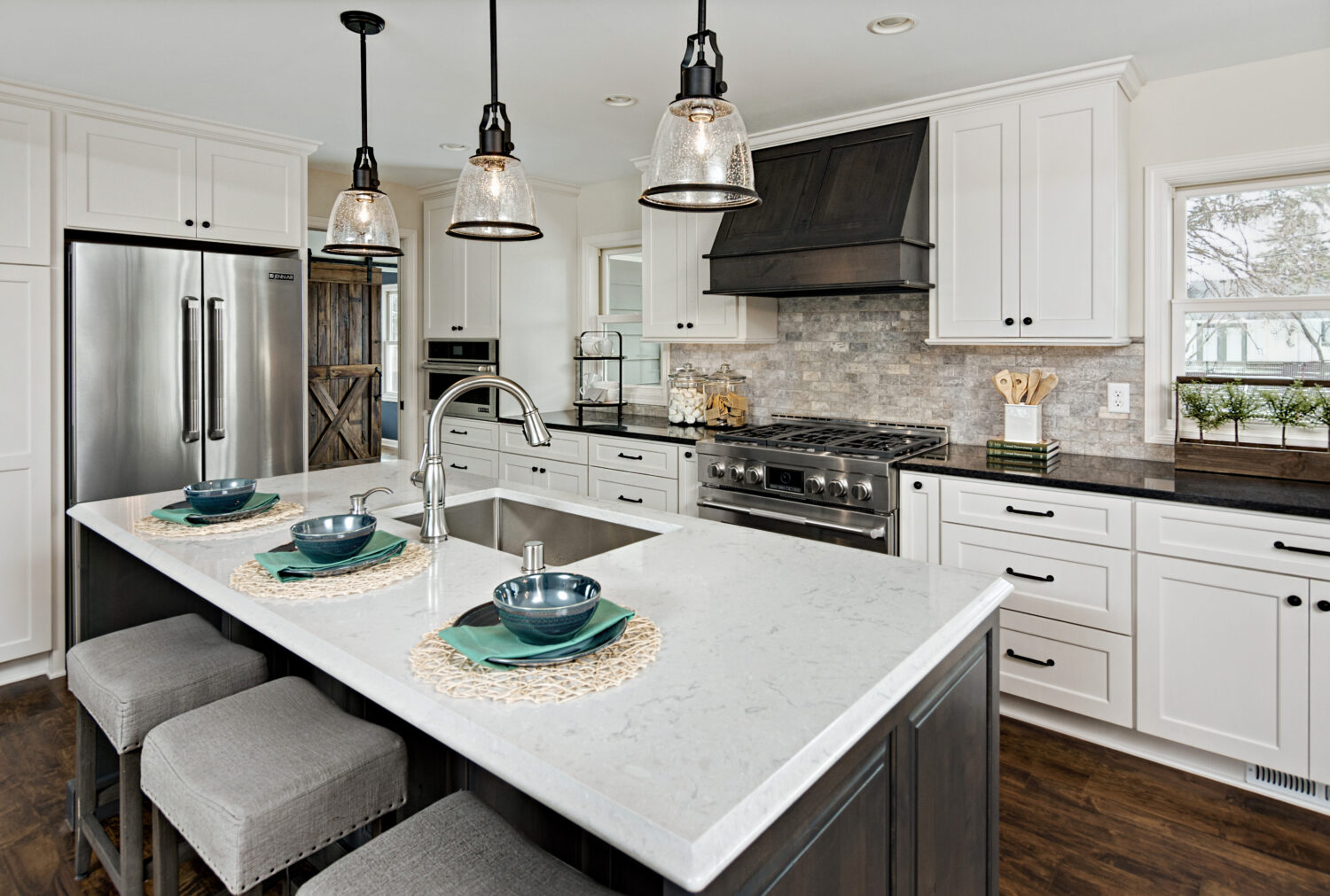 A kitchen island with seating for three with a sink across from the cooktop and gray stained wood hood surrounded by white painted cabinets.