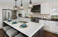 A kitchen island with seating for three with a sink across from the cooktop and gray stained wood hood surrounded by white painted cabinets.