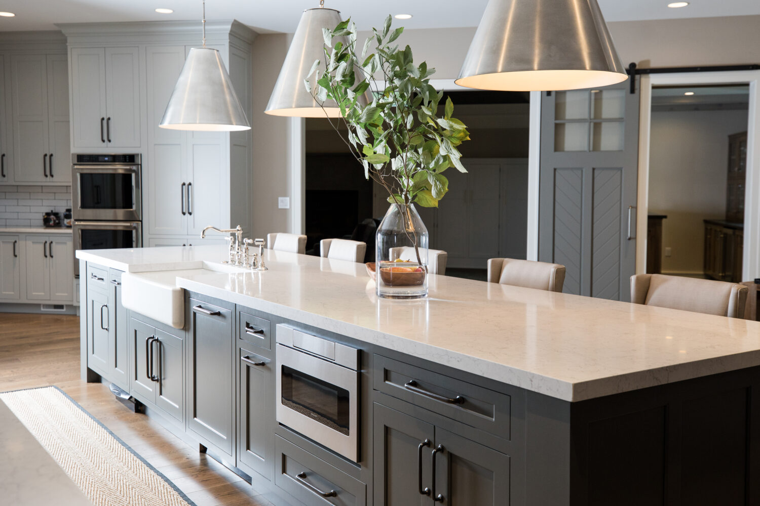 A long and dark gray kitchen island with an apron sink, a microwave drawer, seating for 5, and lots of cabinets storage.