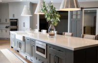 A long and dark gray kitchen island with an apron sink, a microwave drawer, seating for 5, and lots of cabinets storage.