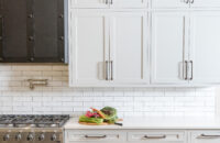 A stunning all white kitchen design with white painted inset shaker cabinets, long dark nickel hardware pulls, white subway tiles, white quartz countertops and a stunning dark metal hood.