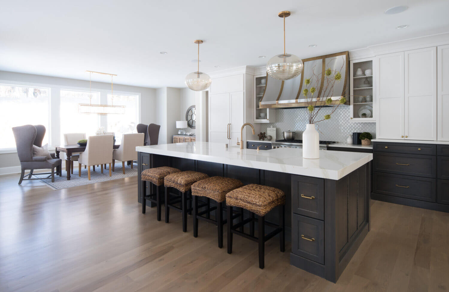A soft black and white kitchen design with dark gray stained oak for the base cabinets and bright white paint for the wall cabinets and appliance panels. Brass hardware, light fixtures, and metal hood are used as accents throughout.