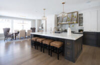 A soft black and white kitchen design with dark gray stained oak for the base cabinets and bright white paint for the wall cabinets and appliance panels. Brass hardware, light fixtures, and metal hood are used as accents throughout.