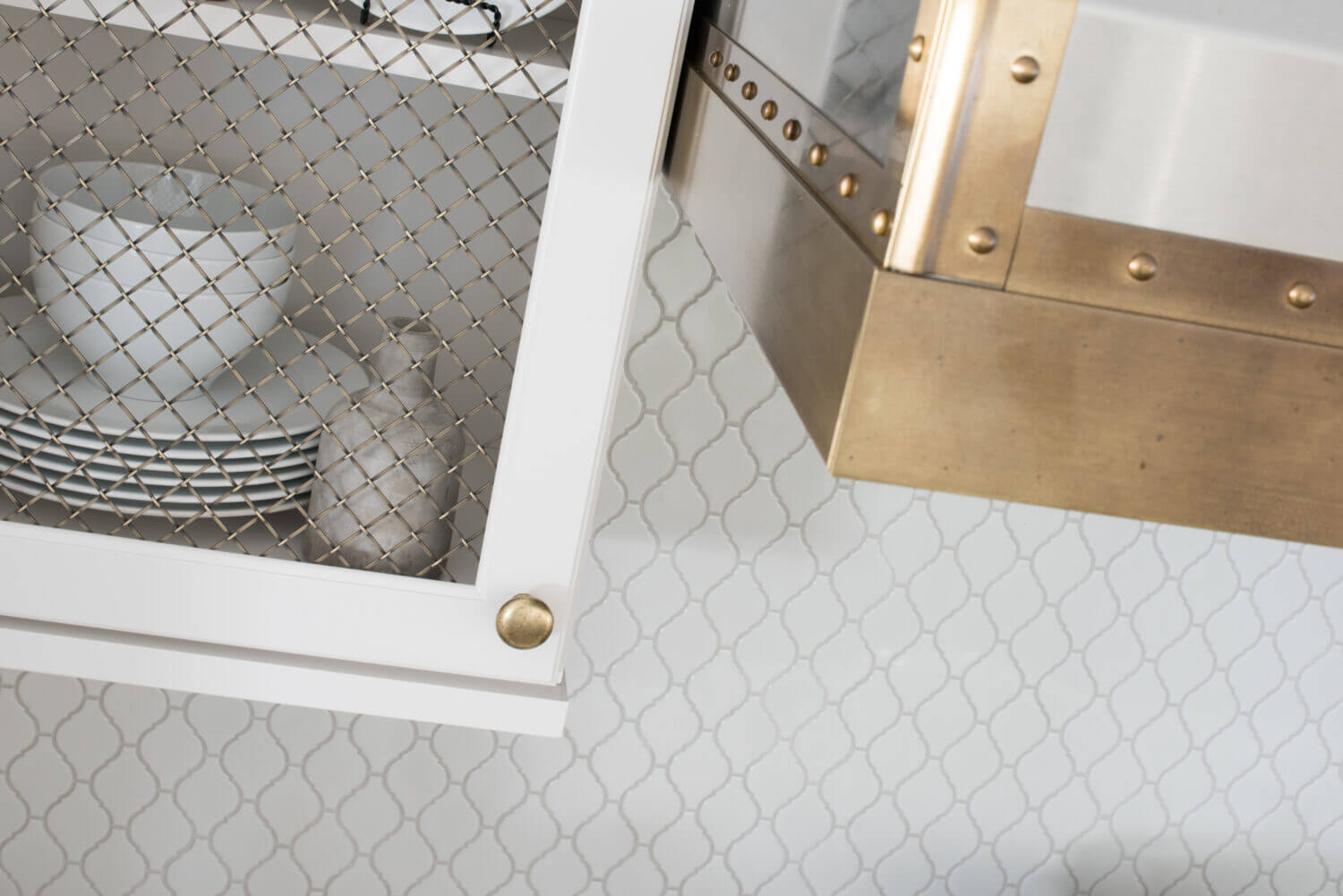 An angled close up of the white painted kitchen wall cabinet with a golden brass mesh metal insert and the coordinating brass metal hood over the cooktop.