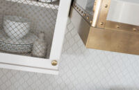 An angled close up of the white painted kitchen wall cabinet with a golden brass mesh metal insert and the coordinating brass metal hood over the cooktop.