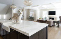 A view over the dark gray stained kitchen island with oak cabinetry into the adjacent living room with a white painted fireplace mantel that coordinated with the White cabinets of the kitchen perimeter.