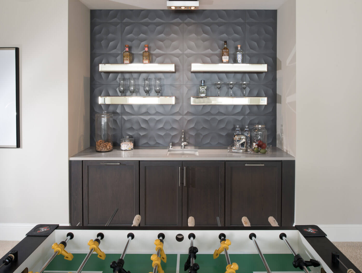 A small nook with dark gray stained cabinets was added in the basement next to the foosball table to store board games, snacks, and supplies for the entertainment room. Stainless steel floating shelves above add space for displaying decor.