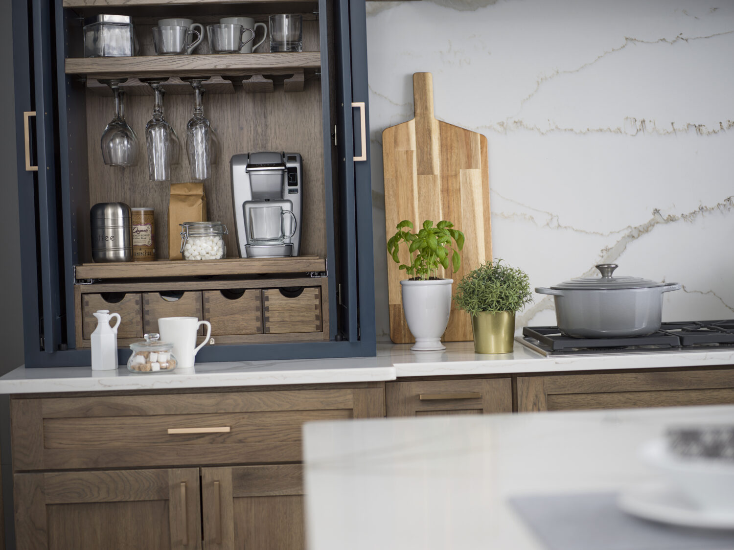 A close up of an open larder cabinet with pocket doors tucked in. Apothecary drawers sit below a flat roll-out shelf with the coffee maker on it. A wine glass rack hangs glasses above.