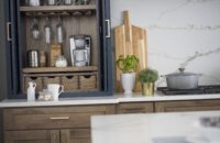 A close up of an open larder cabinet with pocket doors tucked in. Apothecary drawers sit below a flat roll-out shelf with the coffee maker on it. A wine glass rack hangs glasses above.