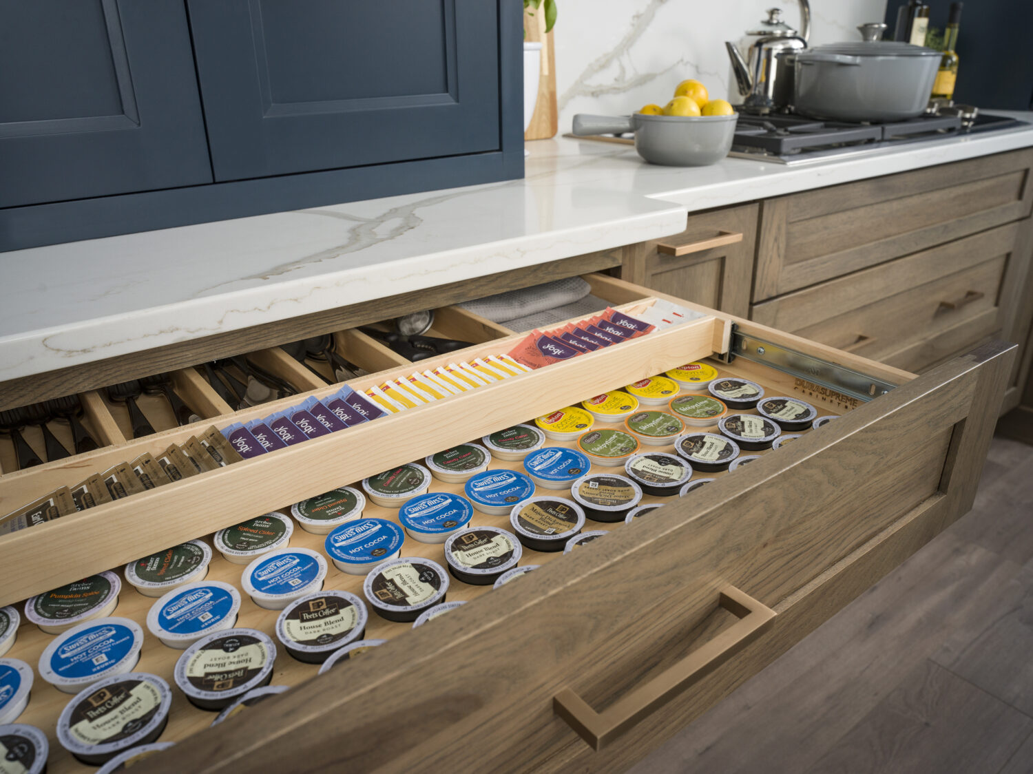 A kitchen drawer opens to show two layers of tiered storage. The top tier has a utensil organizer and storage for tea bags, the bottom is a full drawer K-cup organizer.