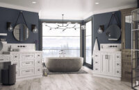 A dream master bathroom with cool navy blue and walls and white painted oak bathroom cabinets and vanities from Dura Supreme. A free standing stone tub sits in front of several large windows with a stunning mountainside view.