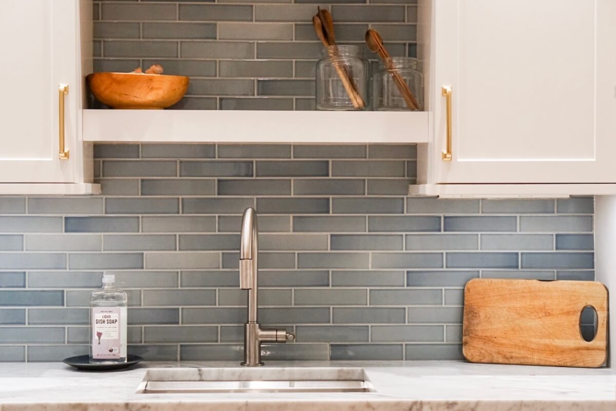 Close up of prep sink area with a floating and open shelf above, white cabinets, gold cabinet pulls, and a pale blue tiled backsplash.