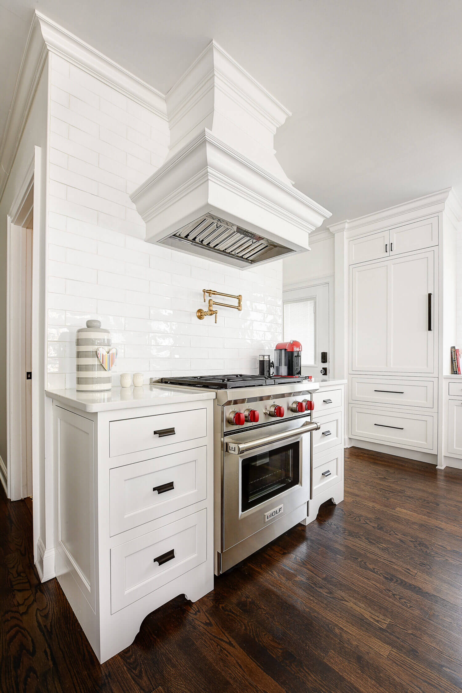 The range an cook top in this remodeled kitchen sits in the center of a the room. The designer highlighted the area with a full wall subway backsplash that surrounds and emphasizes a beautiful white painted wood hood.