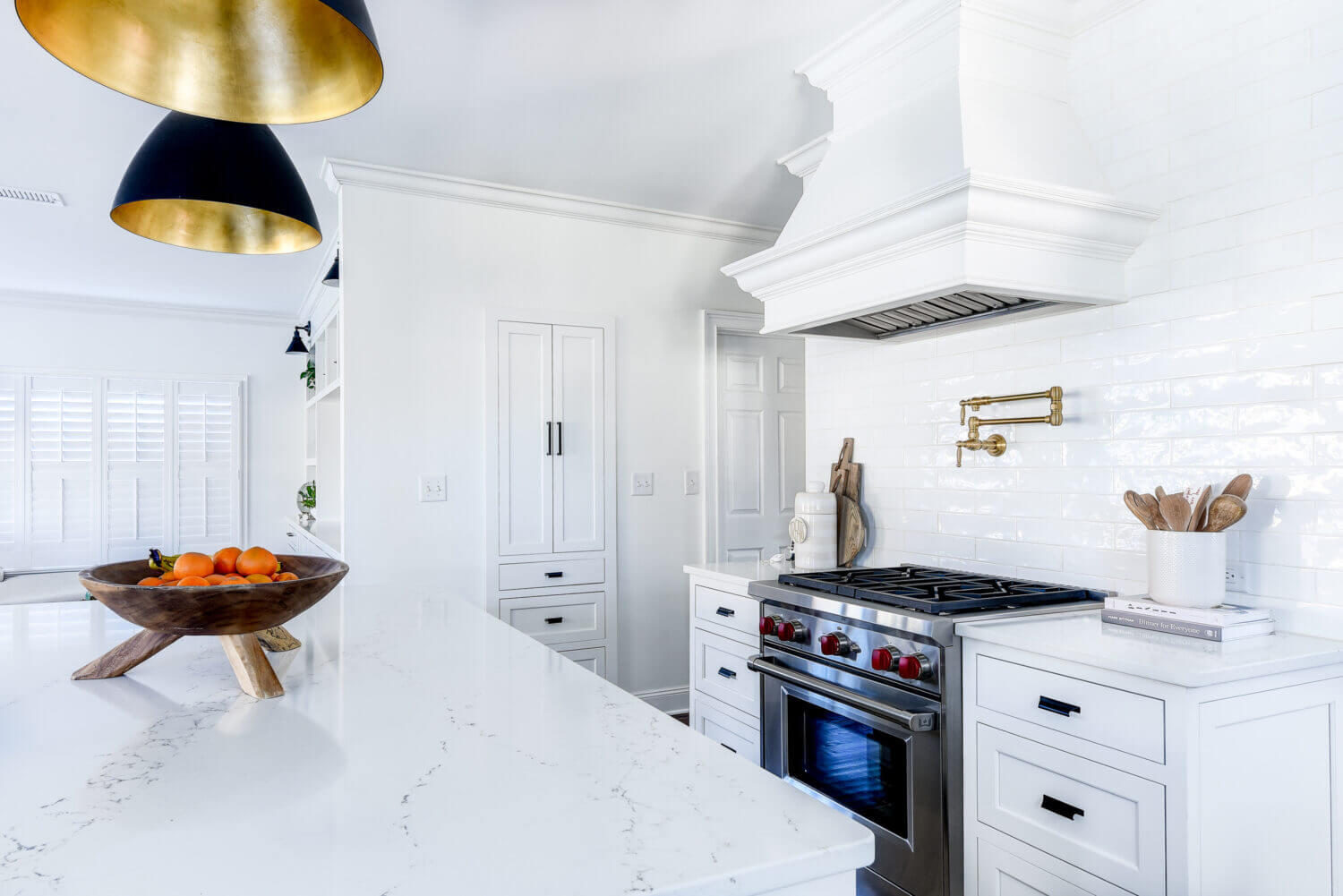 A view over the kitchen island in a white-on-white kitchen design featuring white shaker cabinets, a white independent wood hood, subway tiles, walls, and quartz countertops.