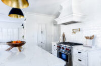 A view over the kitchen island in a white-on-white kitchen design featuring white shaker cabinets, a white independent wood hood, subway tiles, walls, and quartz countertops.