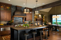 A Rustic Traditional styled kitchen design with a grand wood and metal hood, dark painted kitchen island, and distressed wood cabinets.