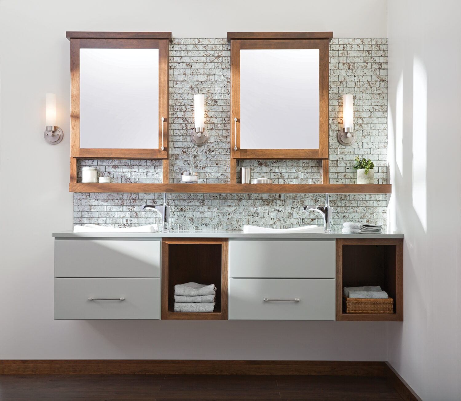 This contemporay bathroom design has a modern style with a floating vanity with dual sets of suspended drawers and two open cabinets for bathroom linens and towels. The double medicine cabinetd with a connecting floating shelf has sleek, streamlined look and adds ti the style of the room.The Dura Supreme cabinetry is shown in a custom matte gray paint accented by medium stain on Cherry wood.