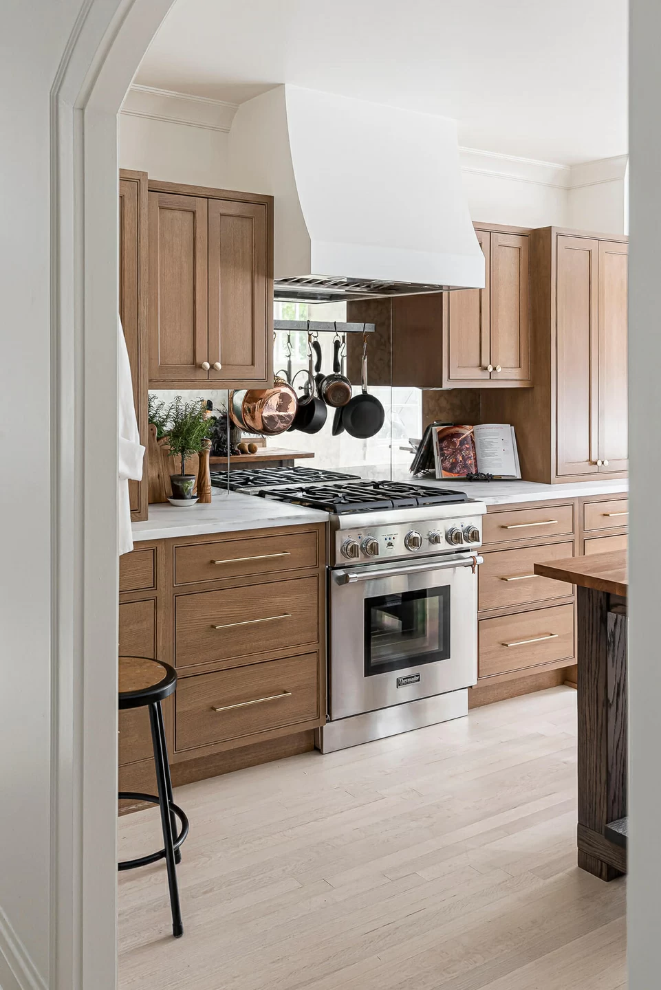 A Dutch style kitchen with American-made inset cabinets in a light stained oak wood. The modern white hood that matches the wall color and a mirror backsplash make this galley kitchen feel larger than it really is.