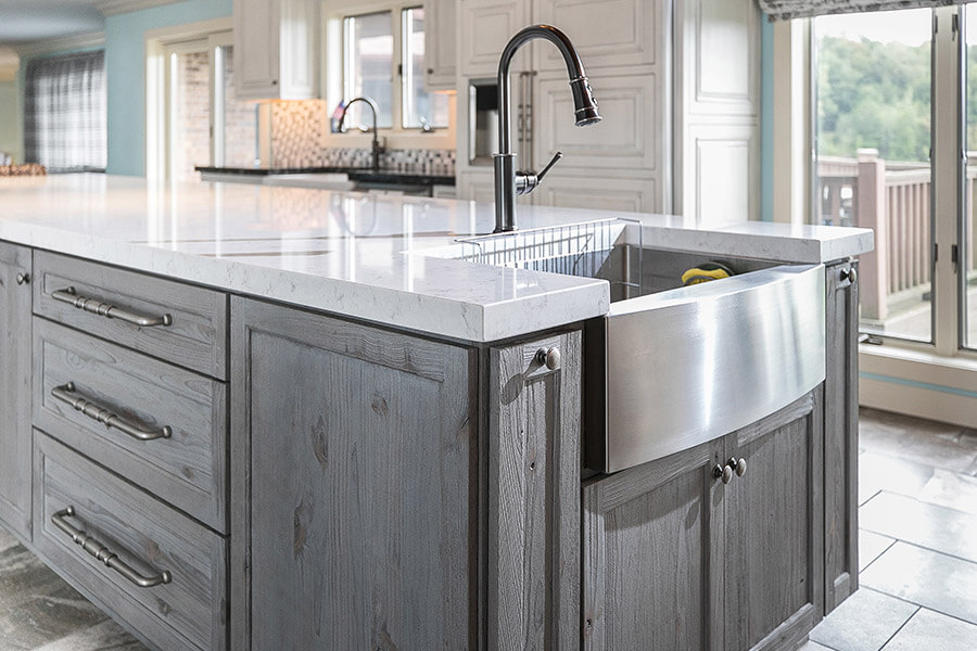 A rustic weathered wood kitchen island with a light gray stain that resembles driftwood. The farmhouse kitchen sink is stainless steel. There is optimal storage surrounding the sink and a long countertop on the island to provide a handy workspace.