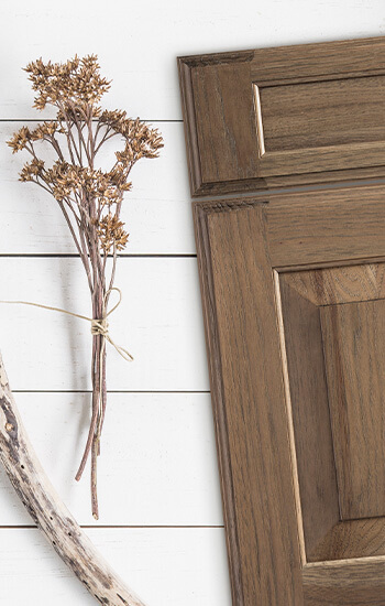 A soft stained cabinet door in Hickory wood with a raised panel door style. The door is laying on top of white painted shiplap. Learn how to make your kitchen design choices for selecting the colors of your cabinetry.