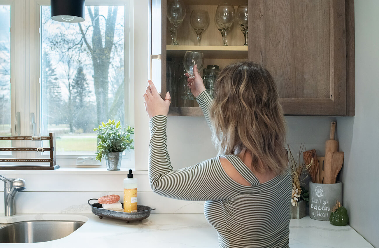 A homeowner in a newly remodeled kitchen taking a glass from a wall cabinet. The medium stained cherry cabinets are solid wood from Dura Supreme Cabinetry. At the start of the design process, take inventory of everything in your kitchen. This includes not only things like cookware and dishes, but features like outlets, recurring paper piles, often used food/drink items, small appliances, and more. This is the perfect time to purge and donate items you no longer need.